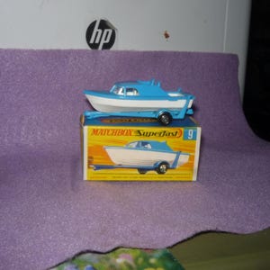 May include: A blue and white toy boat on a matching blue trailer, resting on a yellow and blue cardboard box labeled Matchbox Superfast 9. The boat features a cabin and a small windshield, while the box displays an illustration of the boat.