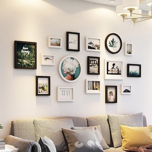 May include: A gallery wall with 15 black and white picture frames of various sizes and shapes. The frames are arranged in a symmetrical pattern on a white wall. The frames contain various images, including a woman with flowers, a landscape, and a cityscape. The text "love living" and "stay" are visible in two of the frames.