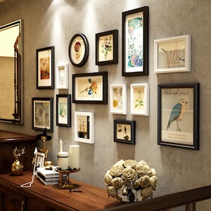 May include: A gallery wall with a variety of framed prints, including botanical illustrations, a bird, and a quote about faith. The frames are in black and white, and the wall is a light gray. There is a wooden table with a vase of white roses in front of the wall.