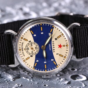 May include: A vintage silver wristwatch with a white face and blue accents. The watch face has black markings and a red star with the word "ПОБЕДА" written in Cyrillic script. The watch has a black nylon strap.
