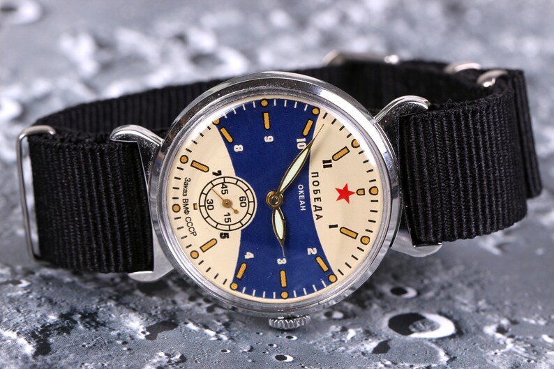 May include: A vintage silver wristwatch with a white face and blue accents. The watch face has black markings and a red star with the word "ПОБЕДА" written in Cyrillic script. The watch has a black nylon strap.