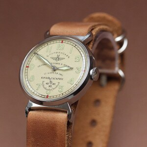 Puede incluir: Un reloj de pulsera plateado con una esfera blanca y agujas negras. La esfera del reloj tiene el texto "12 АПРЕЛЯ 1961 ЮРИЙ ГАГАРИН" y una peque&ntilde;a imagen de un cohete. El reloj est&aacute; en una correa de cuero marr&oacute;n.