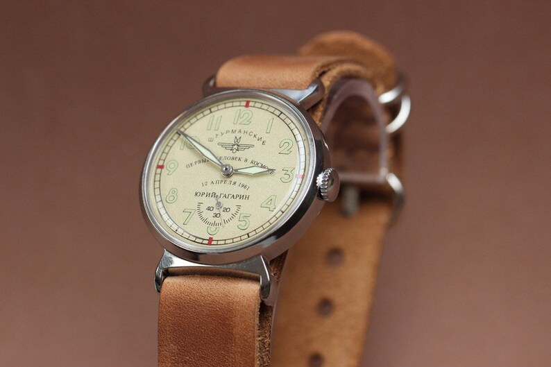 Puede incluir: Un reloj de pulsera plateado con una esfera blanca y agujas negras. La esfera del reloj tiene el texto "12 АПРЕЛЯ 1961 ЮРИЙ ГАГАРИН" y una peque&ntilde;a imagen de un cohete. El reloj est&aacute; en una correa de cuero marr&oacute;n.