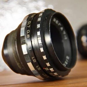 May include: A black camera lens with silver markings. The lens has a depth of field scale marked with numbers from 0.8 to 2. The lens is in focus and the background is blurred.