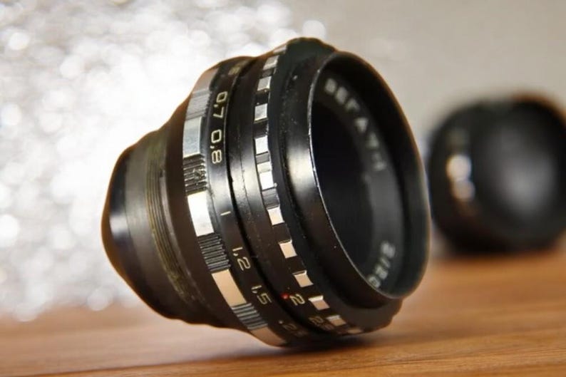 May include: A black camera lens with silver markings. The lens has a depth of field scale marked with numbers from 0.8 to 2. The lens is in focus and the background is blurred.