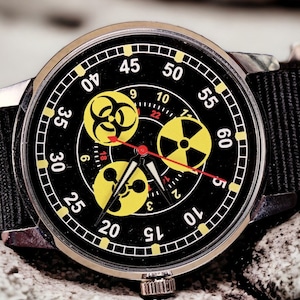 May include: A black wristwatch with a black nylon band. The watch face features yellow hazard symbols and red hands. The watch is set against a textured, gray background.