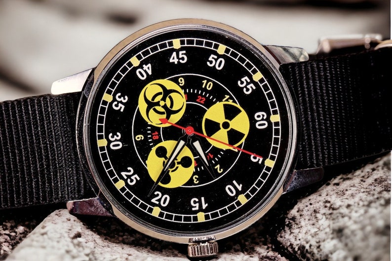 May include: A black wristwatch with a black nylon band. The watch face features yellow hazard symbols and red hands. The watch is set against a textured, gray background.