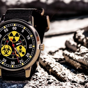 May include: A wristwatch with a black face featuring yellow radiation hazard symbols and white numerals. The watch has a black fabric band and a silver-colored case. The watch hands are red and the watch is set against a blurred, rocky background.