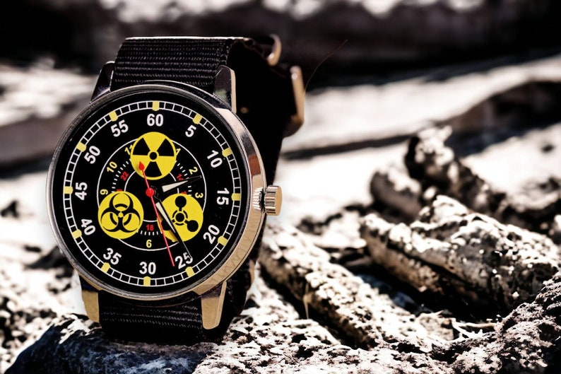 May include: A wristwatch with a black face featuring yellow radiation hazard symbols and white numerals. The watch has a black fabric band and a silver-colored case. The watch hands are red and the watch is set against a blurred, rocky background.