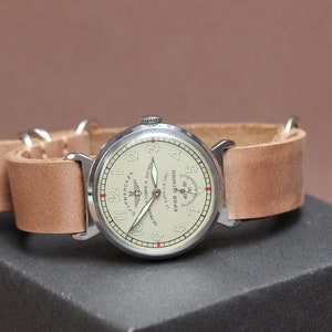 Puede incluir: Un reloj de pulsera plateado con una esfera blanca y agujas negras. El reloj tiene una correa de cuero marr&oacute;n y un peque&ntilde;o logotipo de avi&oacute;n en la esfera. La esfera del reloj tambi&eacute;n tiene el texto "ЧАСЫ АВИАЦИОННЫЕ" en escritura cir&iacute;lica.