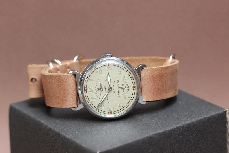 Puede incluir: Un reloj de pulsera plateado con una esfera blanca y agujas negras. El reloj tiene una correa de cuero marr&oacute;n y un peque&ntilde;o logotipo de avi&oacute;n en la esfera. La esfera del reloj tambi&eacute;n tiene el texto "ЧАСЫ АВИАЦИОННЫЕ" en escritura cir&iacute;lica.