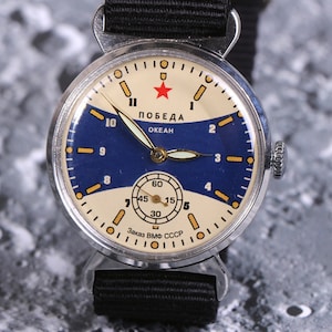 May include: A silver wristwatch with a blue and white face. The watch face has a red star and the text "ПОБЕДА ОКЕАН" in Cyrillic script. The watch strap is black.