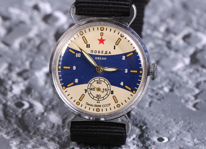 May include: A silver wristwatch with a blue and white face. The watch face has a red star and the text "ПОБЕДА ОКЕАН" in Cyrillic script. The watch strap is black.