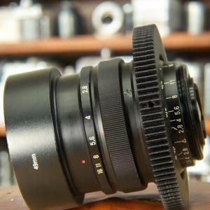 May include: A black camera lens with a gear ring and a 49mm filter thread. The lens has a depth of field scale and aperture markings.