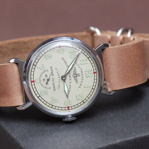 Puede incluir: Un reloj de pulsera plateado con una correa de cuero marr&oacute;n. La esfera del reloj es blanca con n&uacute;meros y manecillas negros. El texto en la esfera del reloj dice "ЮРИЙ ГАГАРИН 12 АПРЕЛЯ 1961 ГОДА СОВЕТСКИЕ ЧАСЫ В КОСМОСЕ ПЕРВЫЙ"