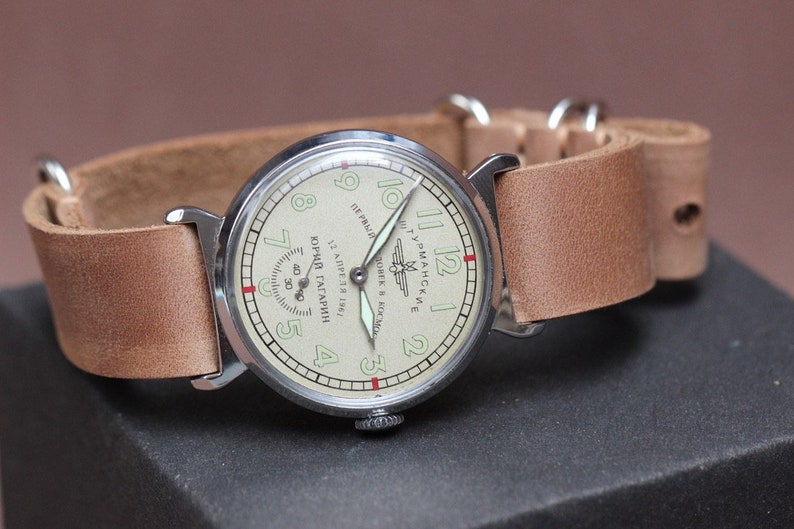 Puede incluir: Un reloj de pulsera plateado con una correa de cuero marr&oacute;n. La esfera del reloj es blanca con n&uacute;meros y manecillas negros. El texto en la esfera del reloj dice "ЮРИЙ ГАГАРИН 12 АПРЕЛЯ 1961 ГОДА СОВЕТСКИЕ ЧАСЫ В КОСМОСЕ ПЕРВЫЙ"