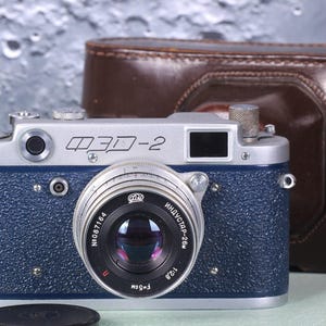 Puede incluir: Cámara plateada y azul vintage con una funda de cuero marrón. La cámara tiene una lente prominente y el texto "ФЭД-2" en la parte delantera. Una tapa de lente está delante de la cámara.