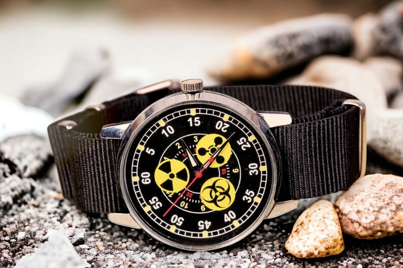 May include: A black wristwatch with a black nylon band. The watch face is black with white numerals and yellow hazard symbols. The watch is set against a background of gray rocks and pebbles.