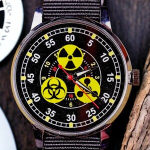May include: A black wristwatch with a silver case and a black fabric band. The watch face features yellow radiation symbols and a red second hand. The numbers are white and yellow.