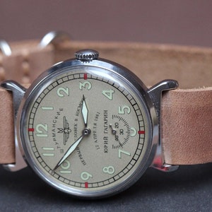 Puede incluir: Un reloj de pulsera plateado con una esfera de color crema y agujas negras. La esfera del reloj tiene marcadores de hora romanos negros y un peque&ntilde;o dial de segundos. La esfera del reloj tambi&eacute;n tiene el texto "ШТУРМАНСКИЕ" y "ПЕРВЫЙ ЧЕЛОВЕК В КОСМОС 12 АПРЕЛЯ 1961 ЮРИЙ ГАГАРИН" en escritura cir&iacute;lica.