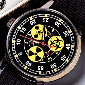 May include: A black wristwatch with a black nylon band. The watch face is black with yellow and white markings, including three yellow hazard symbols. The watch hands are red. The watch has a stainless steel case.