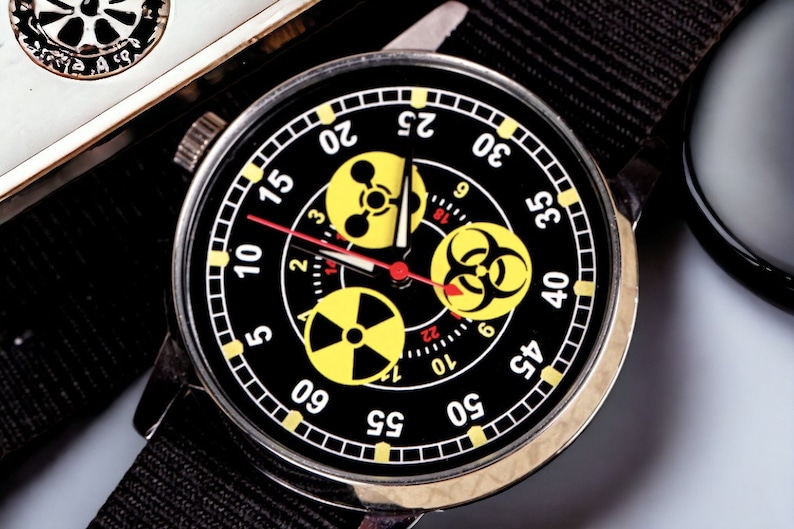 May include: A black wristwatch with a black nylon band. The watch face is black with yellow and white markings, including three yellow hazard symbols. The watch hands are red. The watch has a stainless steel case.