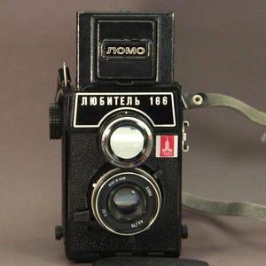 May include: A black LOMO Lubitel 166 camera with a leather strap. The camera has a silver lens and a red and white sticker with the Olympic rings and a tower.
