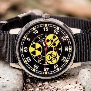 May include: A black wristwatch with a black nylon band. The watch face is black with white numerals and yellow hazard symbols. The watch is set against a background of rocks.