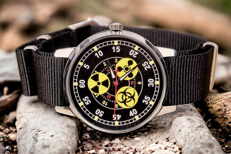 May include: A black wristwatch with a black nylon band. The watch face is black with white numerals and yellow hazard symbols. The watch is set against a background of rocks.