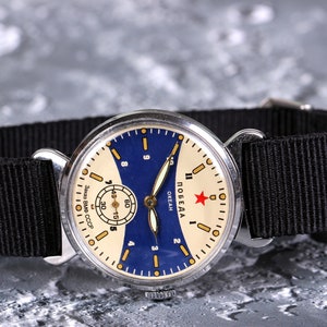 May include: A vintage silver wristwatch with a white face and blue accents. The watch face has black hands and a red star with the word "ПОБЕДА" and "OKEAH" printed on it. The watch is on a black nylon strap.