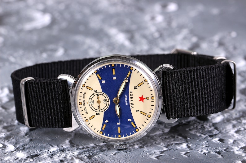 May include: A vintage silver wristwatch with a white face and blue accents. The watch face has black hands and a red star with the word "ПОБЕДА" and "OKEAH" printed on it. The watch is on a black nylon strap.