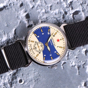 May include: A vintage wristwatch with a white face and blue accents. The watch has a black nylon strap and the words "Победа Океан" (Pobeda Ocean) printed on the face.