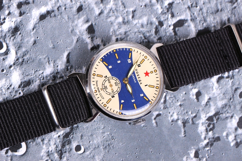May include: A vintage wristwatch with a white face and blue accents. The watch has a black nylon strap and the words "Победа Океан" (Pobeda Ocean) printed on the face.