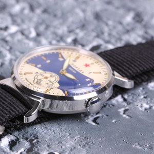 May include: A silver wristwatch with a white face and blue accents. The watch face features a red star and the word "Soviet" in Cyrillic script. The watch is on a black nylon strap.
