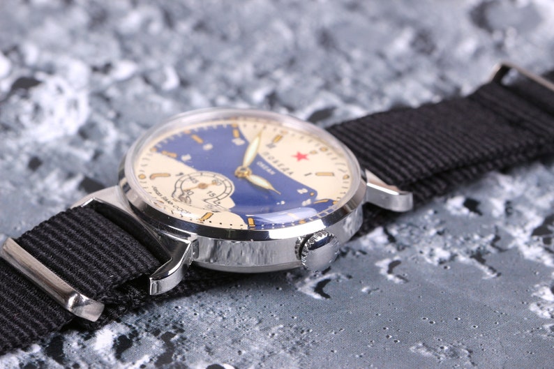 May include: A silver wristwatch with a white face and blue accents. The watch face features a red star and the word "Soviet" in Cyrillic script. The watch is on a black nylon strap.