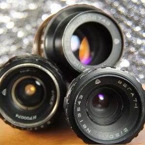 May include: Three vintage camera lenses with black metal bodies and silver markings. The lenses are arranged in a triangular shape on a wooden surface.