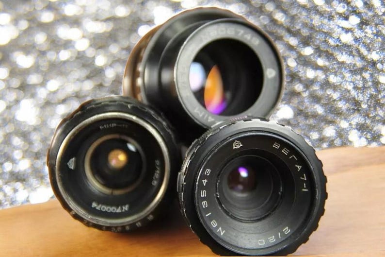 May include: Three vintage camera lenses with black metal bodies and silver markings. The lenses are arranged in a triangular shape on a wooden surface.