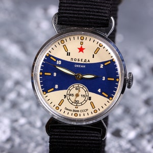 May include: A vintage silver wristwatch with a blue and white face. The watch face has a red star and the text "ПОБЕДА ОКЕАН" and "Заказ ВМФ СССР".