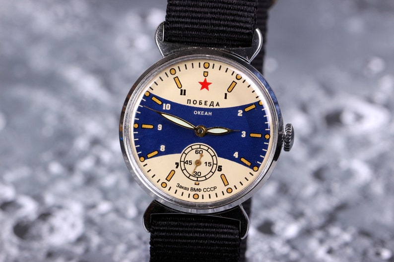 May include: A vintage silver wristwatch with a blue and white face. The watch face has a red star and the text "ПОБЕДА ОКЕАН" and "Заказ ВМФ СССР".