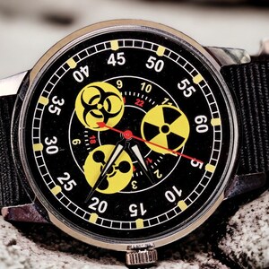 May include: A black wristwatch with a black nylon band. The watch face features yellow hazard symbols and red hands. The watch is set against a textured, gray background.