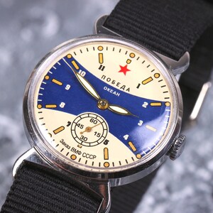 May include: A vintage silver wristwatch with a white face and blue accents. The watch face features the text "ПОБЕДА ОКЕАН" and a red star. The watch strap is black.