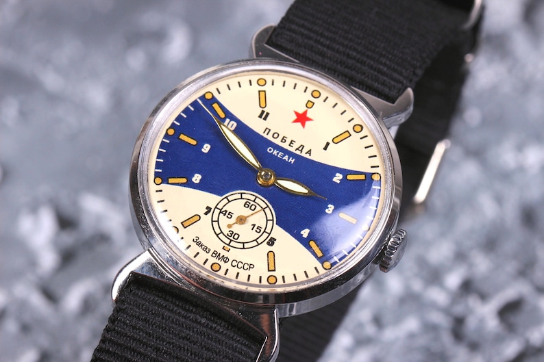 May include: A vintage silver wristwatch with a white face and blue accents. The watch face features the text "ПОБЕДА ОКЕАН" and a red star. The watch strap is black.