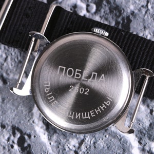 May include: The back of a silver wristwatch with a black nylon strap. The watch face has the word "Победа" and the number "2602" engraved on it. The text "Пыле" and "Чищенны" are also engraved on the watch face.