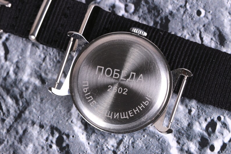 May include: The back of a silver wristwatch with a black nylon strap. The watch face has the word "Победа" and the number "2602" engraved on it. The text "Пыле" and "Чищенны" are also engraved on the watch face.