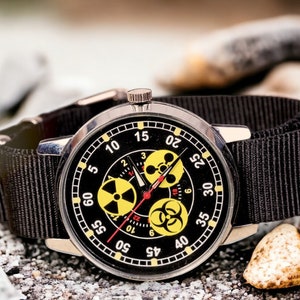 May include: A black wristwatch with a black nylon band. The watch face is black with white numerals and yellow hazard symbols. The watch is set against a background of gray rocks and pebbles.