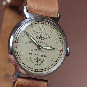 Puede incluir: Un reloj de pulsera vintage de plata con una esfera de color crema. La esfera presenta marcadores de hora romanos negros, agujas negras y un peque&ntilde;o dial de segundos. El reloj tiene una correa de cuero marr&oacute;n y el texto "ШТУРМАНСКИЕ ПЕРВЫЙ ЧЕЛОВЕК В КОСМОС 12 АПРЕЛЯ 1961 ЮРИЙ ГАГАРИН" est&aacute; impreso en la esfera.