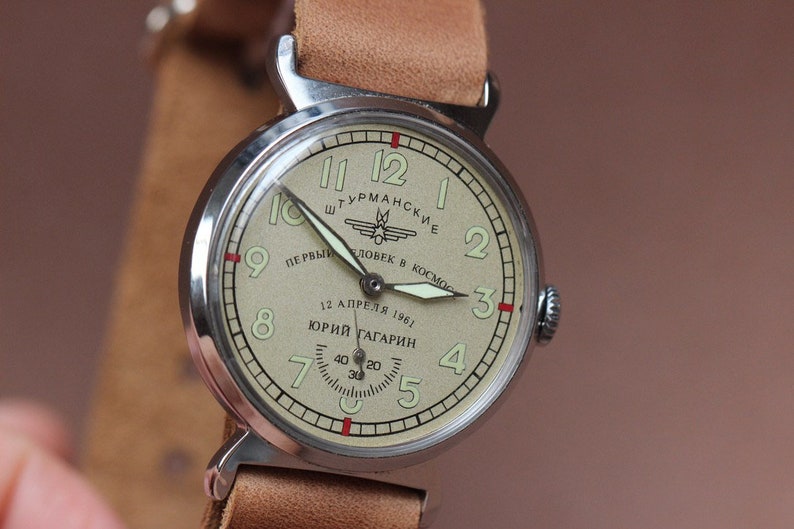Puede incluir: Un reloj de pulsera vintage de plata con una esfera de color crema. La esfera presenta marcadores de hora romanos negros, agujas negras y un peque&ntilde;o dial de segundos. El reloj tiene una correa de cuero marr&oacute;n y el texto "ШТУРМАНСКИЕ ПЕРВЫЙ ЧЕЛОВЕК В КОСМОС 12 АПРЕЛЯ 1961 ЮРИЙ ГАГАРИН" est&aacute; impreso en la esfera.