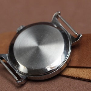 Puede incluir: La parte posterior de un reloj de pulsera plateado con una caja de metal lisa y pulida. El reloj est&aacute; sujeto a una correa de cuero marr&oacute;n.