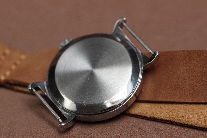 Puede incluir: La parte posterior de un reloj de pulsera plateado con una caja de metal lisa y pulida. El reloj est&aacute; sujeto a una correa de cuero marr&oacute;n.