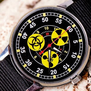 May include: A black wristwatch with a black nylon band. The watch face features yellow hazard symbols and white numerals against a black background. The watch has a silver-colored metal case.
